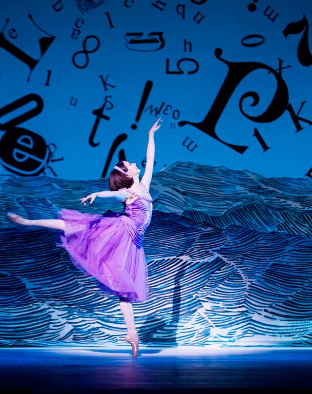 Alice in a lilac dress balancing on one pointe, in front of a backdrop of floating letters and waves