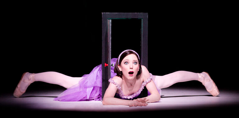 Alice in a lilac dress lying in the splits with her head through a small door, looking startled