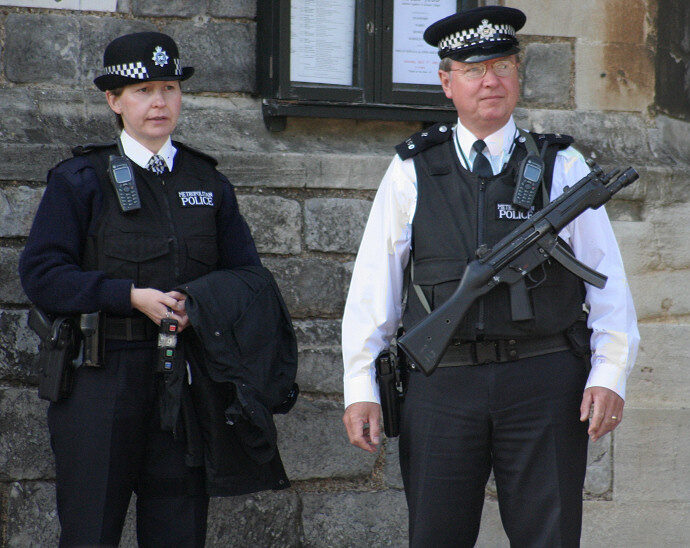 Two Metropolitan Police officers in chequered caps and black protective vests standing by a stone wall, one of them holding a submachine gun