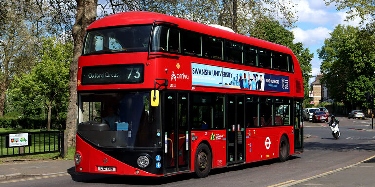 A red New Routemaster double-decker bus with "Oxford Circus 73" on its front sign, turning on a sunny tree-lined street
