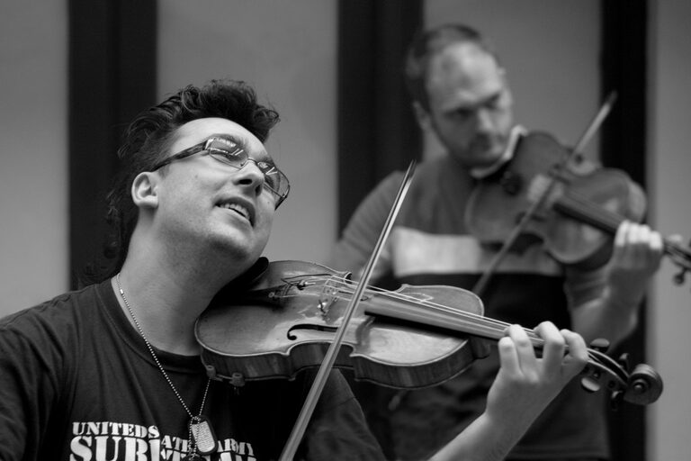 Two men playing violins, the one in front wearing glasses with his head tilted back, in black and white