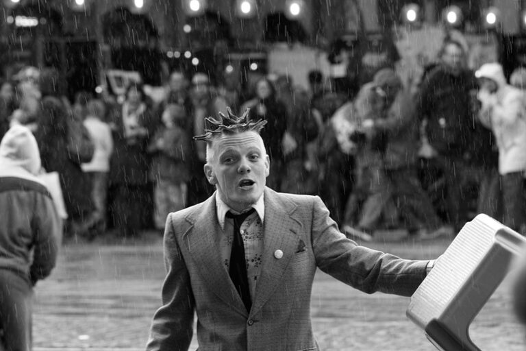 A street performer with spiked hair, in a tweed jacket and tie, calling out to the crowd in the rain, in black and white
