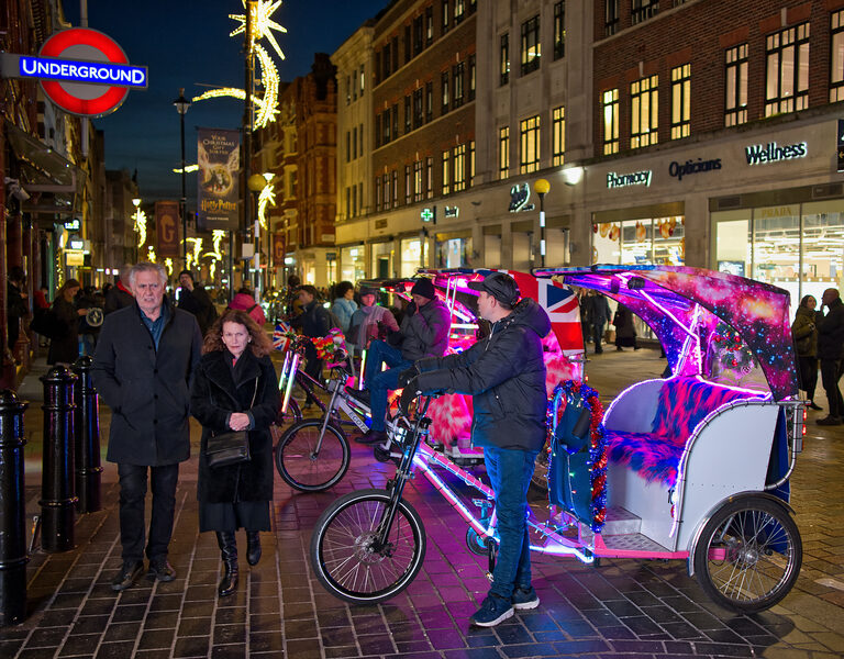 A line of rickshaws decorated with pink lights, fake fur and Union Jack flags waiting on a shopping street at night, as a couple walk past