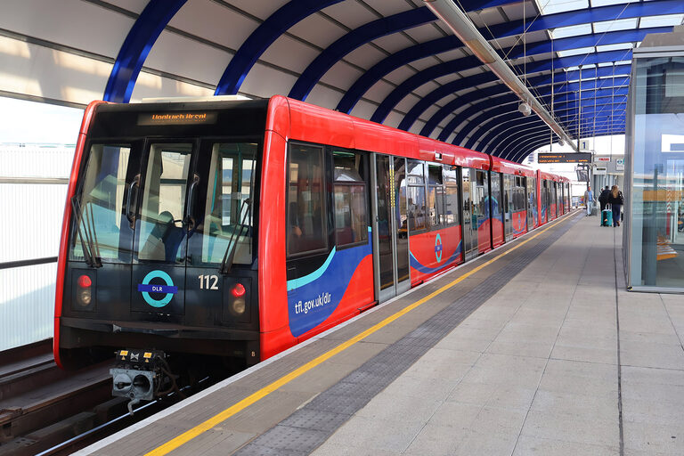 A red and blue DLR train numbered 112 waiting at London City Airport station, under a curved roof with blue ribs, with two travellers and a suitcase far down the platform