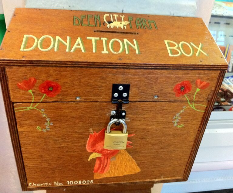 A wooden box painted with red poppies and a cockerel, lettered "Deen City Farm Donation Box" across the top, with a padlock on the front