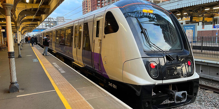 A white and purple Elizabeth line train waiting at a platform at Southall station, under an old ironwork canopy, with a few passengers further along