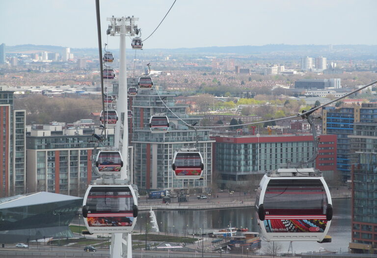 Red and white cable cars coming in to the Royal Docks, with a pylon beside them and the glass Crystal building, Royal Victoria Dock and blocks of flats below