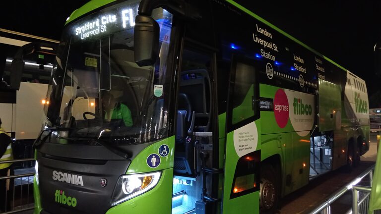 The front of a bright green Flibco coach with its door open at the Stansted Airport coach station at night, with "Stratford City, Liverpool St" on its sign