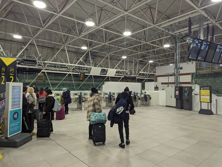 Travellers with suitcases at the ticket gates of Gatwick Airport station, beside a sign saying you can pay for trains to London with Oyster or contactless at the gates