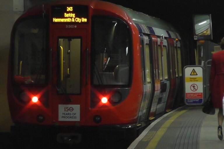 The red front of a Tube train at a dimly lit underground platform, the screen on its front reading Barking, Hammersmith & City line