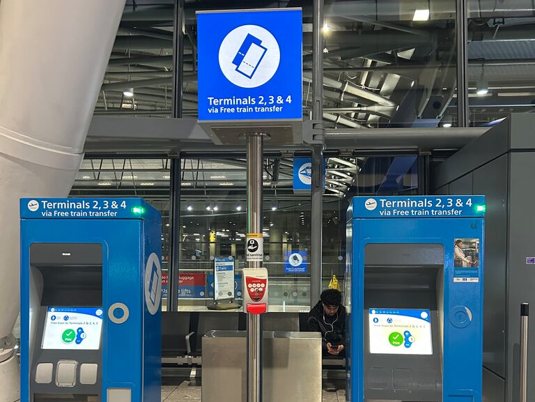 Two blue ticket machines at Heathrow Terminal 5, under a sign reading "Terminals 2, 3 & 4 via Free train transfer"