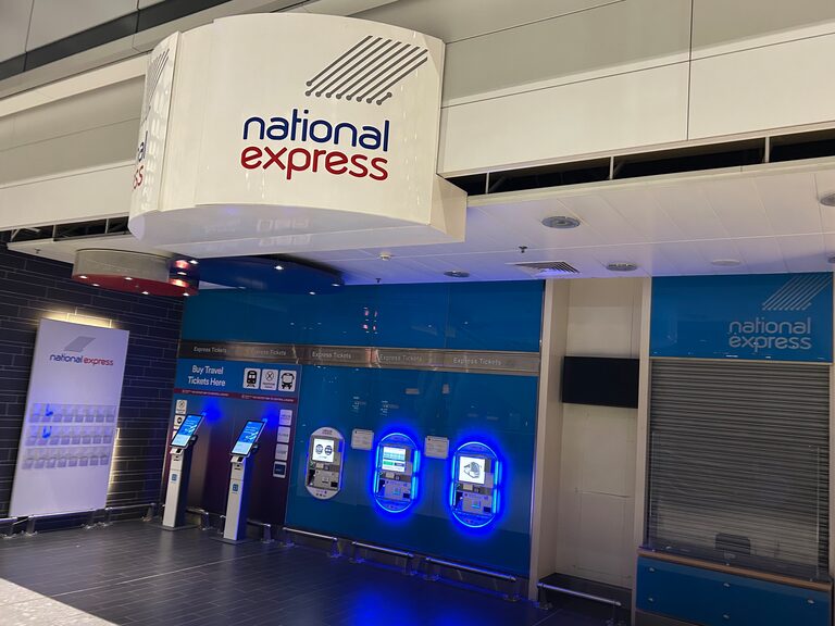 Self-service ticket machines lit in blue under a round National Express sign at Heathrow Terminal 5, beside a ticket desk with its shutter down