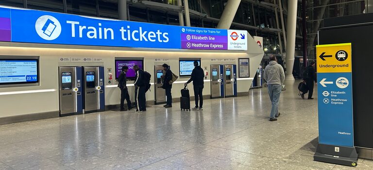 A row of silver and blue ticket machines under a long blue "Train tickets" sign at Heathrow Terminal 5, with a few travellers using them and a sign on the right pointing to the Underground, the Elizabeth line and the Heathrow Express