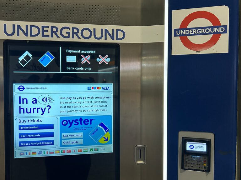 A London Underground ticket machine with a touch screen offering tickets and Oyster cards, a row of small flags along the bottom of the screen, a card reader to its right and an Underground roundel above
