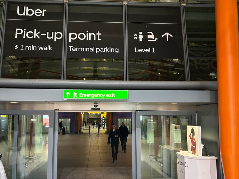 A large sign above a terminal exit at Heathrow Terminal 5 reading "Uber Pick-up point, 1 min walk, Terminal parking, Level 1", with people walking through the doors below
