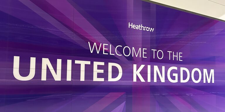 A long purple wall at Heathrow Terminal 5 reading "Heathrow" and "Welcome to the United Kingdom" in white letters, over a faint Union Jack pattern