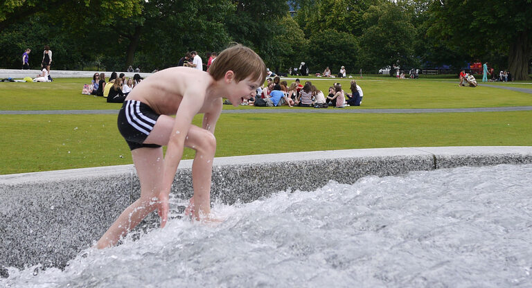 A boy in swimming trunks wading up through fast-flowing water in the Diana Memorial Fountain, with picnickers on the grass behind