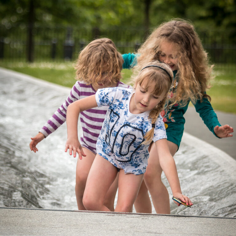 Three girls wading together through the shallow, rippling water of the Diana Memorial Fountain