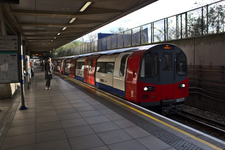A red, white and blue Jubilee line Tube train pulling into the platform at Canning Town station, with a few people on the platform