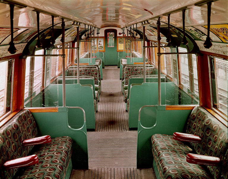Inside an old Tube carriage, with green and cream panels, patterned seats with red armrests and straps hanging from the ceiling