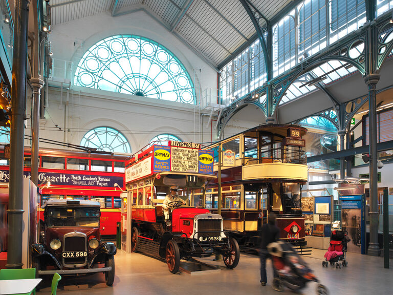 Old double-decker buses and an old black taxi lined up under the museum's iron and glass roof