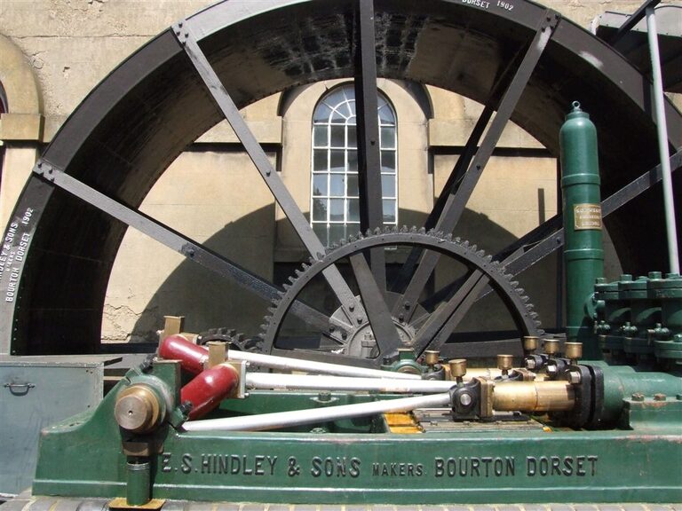 A green three-cylinder water pump with brass fittings, marked "E. S. Hindley & Sons, makers, Bourton, Dorset", in front of the large iron water wheel that drives it
