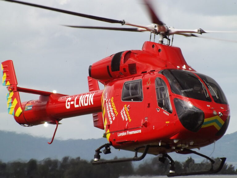 A red helicopter with yellow and green stripes, marked "G-LNDN" and "London's Air Ambulance", flying just above the ground