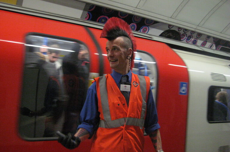 A London Underground worker in an orange hi-vis vest, with a tall red mohican, standing on a platform beside a red and white train