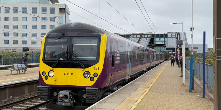 A purple East Midlands Railway train with a yellow front and Luton Airport Express lettering by the cab, at a platform at Luton Airport Parkway station