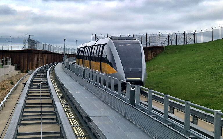A silver and gold driverless Luton DART shuttle car coming along its track in a cutting, beside a grassy bank and a tall security fence