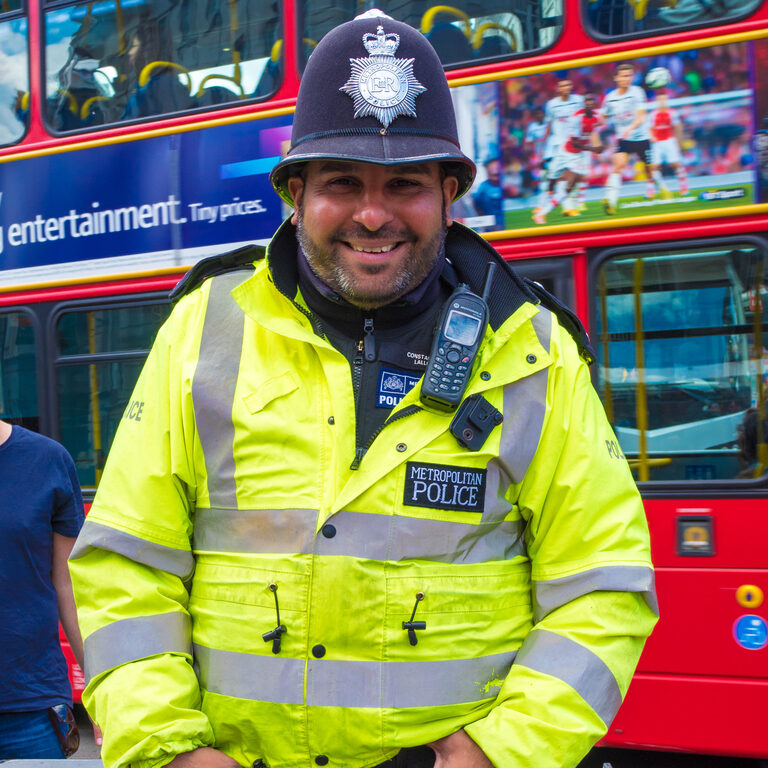 A smiling police officer in a black helmet with a silver star badge and a yellow high-visibility jacket marked "Metropolitan Police", with a red double-decker bus behind him