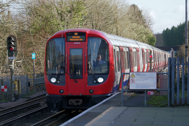 A red-fronted Tube train with its headlights on at an open-air platform beside bare trees, the screen on its front reading Amersham, Metropolitan line