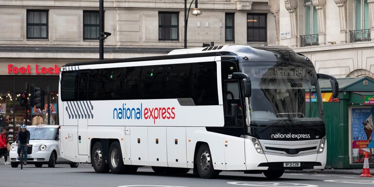 A white National Express coach with blue and red lettering on Park Lane in London, with a white taxi and a cyclist behind it