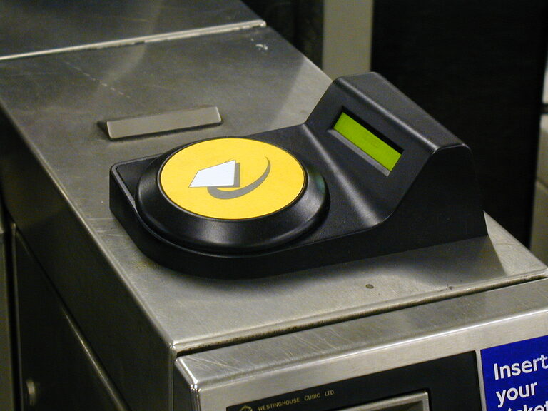 A yellow card reader with the Oyster logo on top of a London Underground ticket gate, next to a small green screen