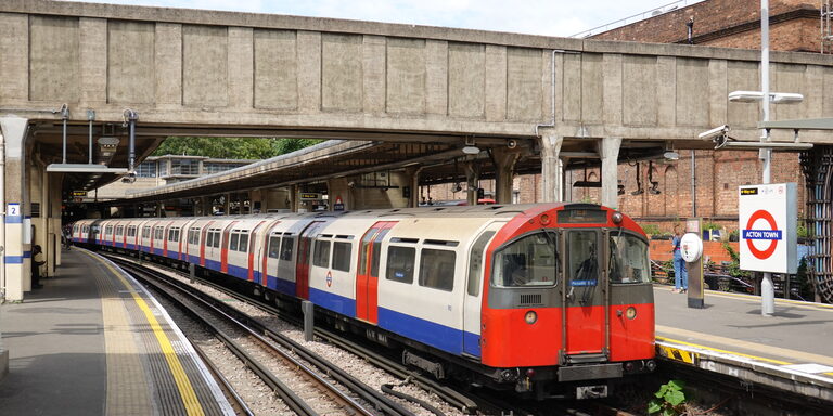 A red, white and blue Piccadilly line Tube train at an open-air platform at Acton Town station, next to a round Acton Town station sign
