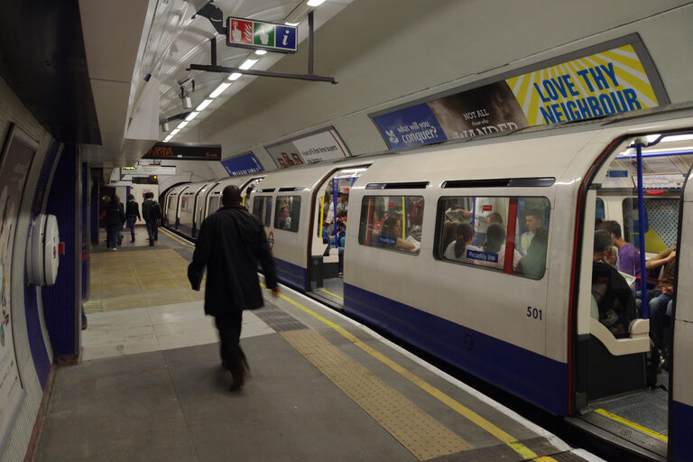 A white, blue and red Tube train with its doors open and Piccadilly line stickers on its windows, as a man walks along the platform
