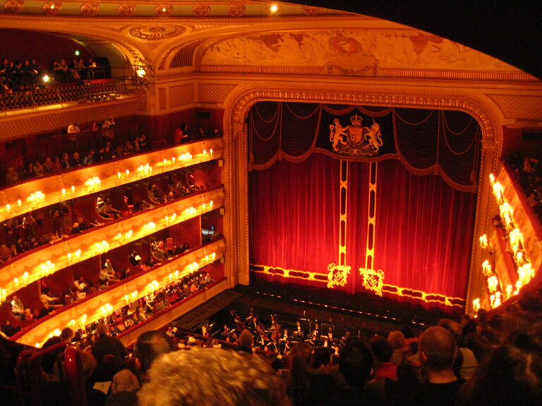 The view down to the red stage curtain with the royal coat of arms, past tiers of lit boxes and the orchestra pit