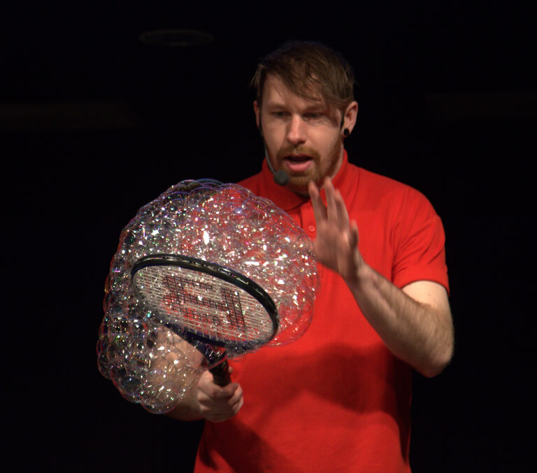A presenter in a red shirt holding up a tennis racket covered in a cluster of shimmering bubbles