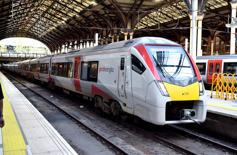 A white Greater Anglia train with a yellow nose and red stripes, used on the Stansted Express, at a platform under the iron roof of London Liverpool Street station