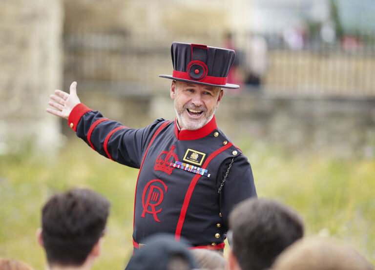 A smiling Yeoman Warder in a dark blue and red uniform and hat, with one arm held out, talking to a group of visitors