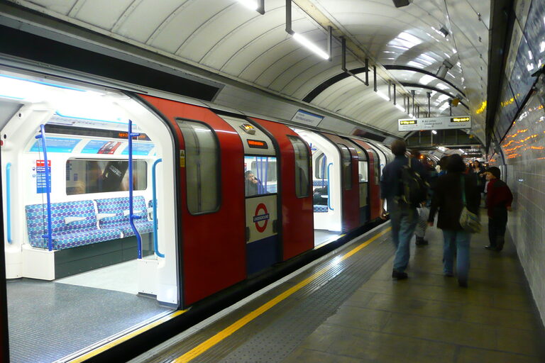 A Tube train with its red doors open at a curved underground platform, showing the blue patterned seats inside
