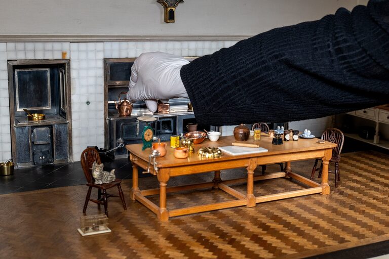 A gloved hand reaching into a miniature kitchen with a black range, a copper kettle, a wooden table laid with pans and jars, and a toy cat on a chair