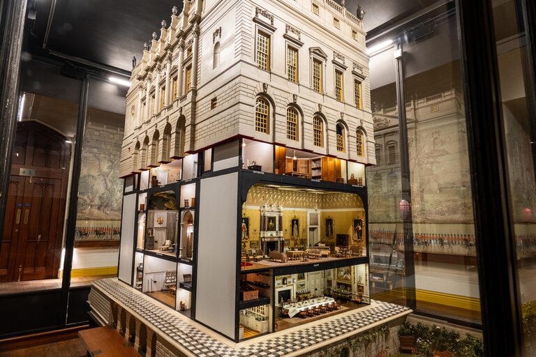 A large model house in a glass case, its upper floors a stone front with rows of arched windows, its lower floors open to show furnished, lit rooms