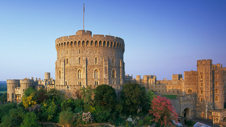 The round stone Round Tower with a flag on top, standing on a mound of trees and flowers, with the castle's walls and square towers on either side under a blue sky