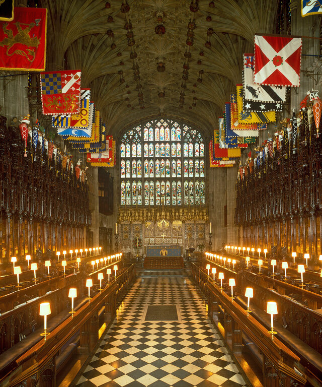 Rows of carved wooden stalls lit by small lamps under colourful hanging banners, with a black-and-white chequered floor leading to a tall stained-glass window