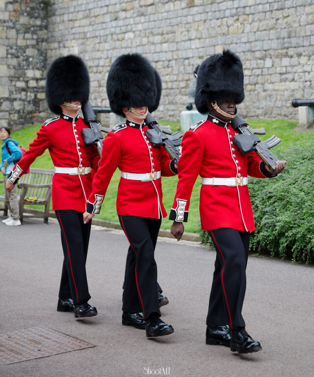 Three guards in red tunics, black trousers and tall black bearskin hats marching along a path, with rifles on their shoulders, past a grass bank and a high stone wall