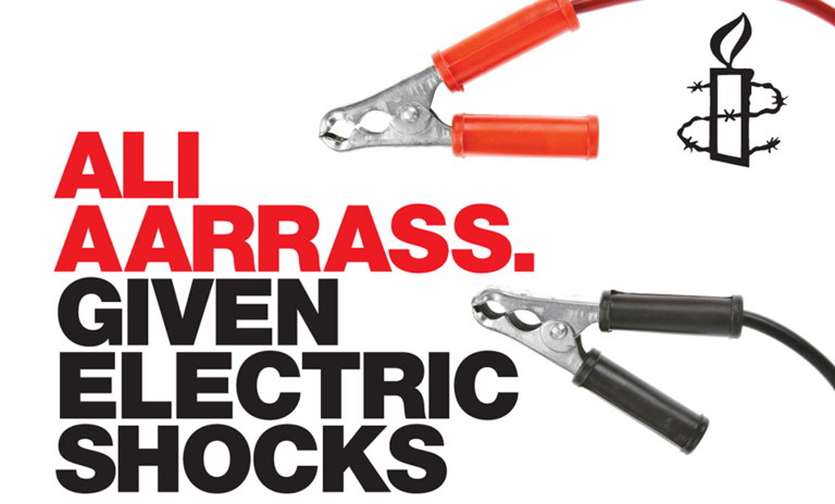 An Amnesty poster reading "Ali Aarrass. Given electric shocks.", with a pair of jump leads and a candle wrapped in barbed wire