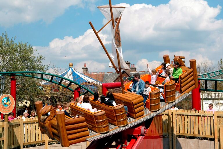 A small pirate ship ride with wooden-log seats tipped up at an angle, full of children and parents, some of them waving