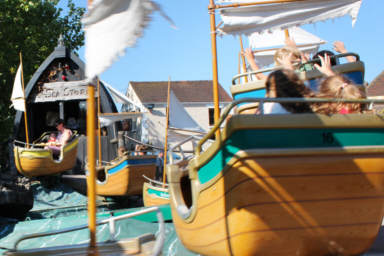 Children in small wooden boats with white sails going round a ride, some of them with their arms in the air