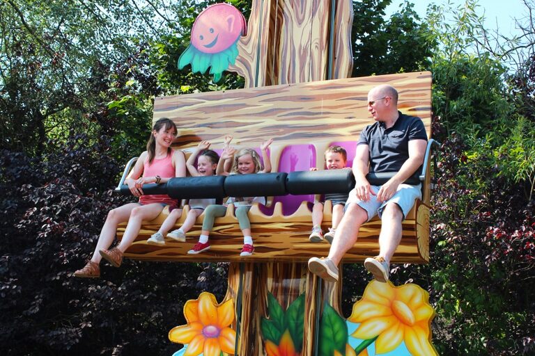 Two adults and three young children sitting in a row on a log-shaped seat high up a cartoon tree trunk, two of the children waving
